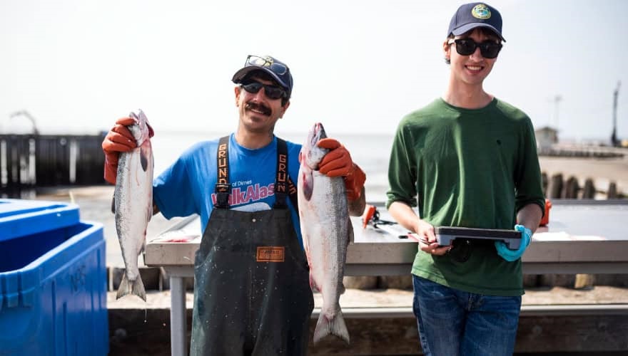 Two men in caps and sunglasses, one holding freshly caught salmon, one with tablet computer